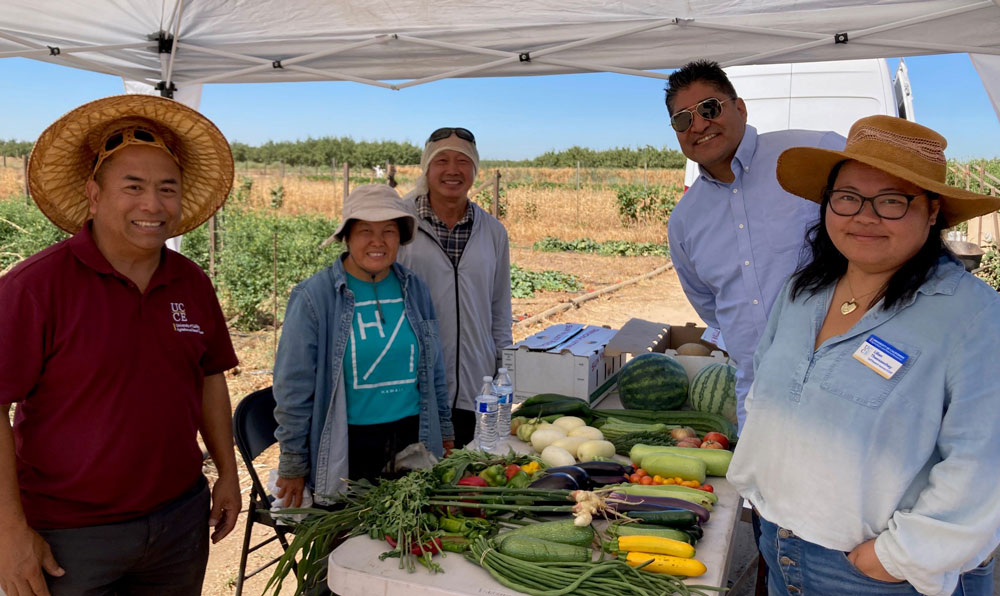 Five people stand around a table of a variety of vegetables with a farm in the background.